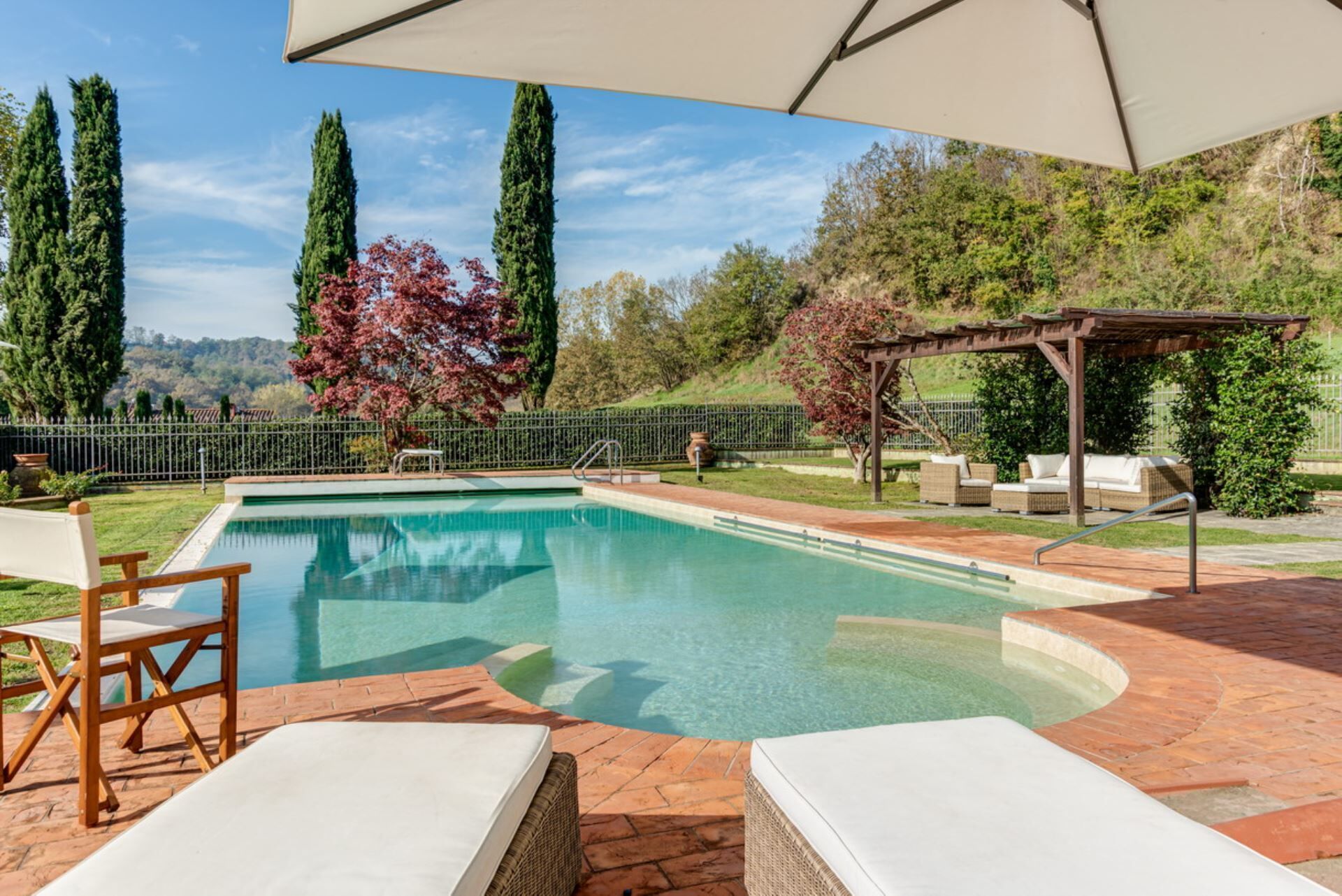 Une piscine dotée de chaises longues et d’une pergola, entourée d’un jardin clos planté d’arbres, offrant une vue sur les collines.
