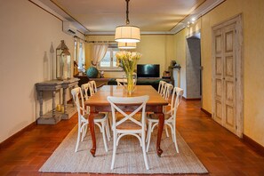 Un comedor con una mesa de madera, sillas blancas, un jarrón con flores, un televisor y una lámpara decorativa.