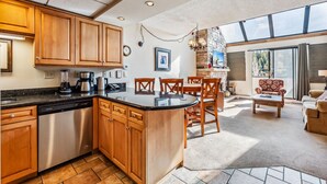 A spacious kitchen with wooden cabinets, a dishwasher, and a large island. There is a dining area with a table and chairs, and a view of the outdoors through large windows. - Breck Ski-In/Ski-Out Condo with Resort Amenities (Breckenridge)