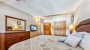 A bedroom with a large bed, wooden dresser, and a mirror. There is a door leading to another room and a wall-mounted TV. - Breck Ski-In/Ski-Out Condo with Resort Amenities (Breckenridge)