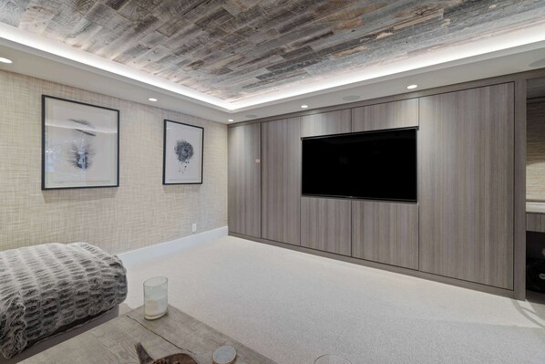 Un dormitorio moderno con un televisor de pantalla plana grande, paneles de madera en las paredes, una cama con manta estampada y cuadros en la pared.
