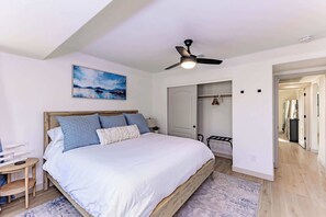 A bedroom with a bed, a ceiling fan, a painting, a closet, and a door leading to another room.