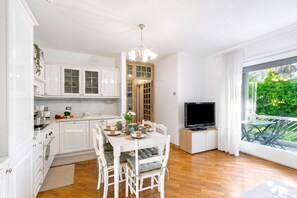 A modern kitchen with white cabinetry, a dining table with chairs, a television, and a large window with a view of greenery. - Riva's Garden Home (Riva del Garda)
