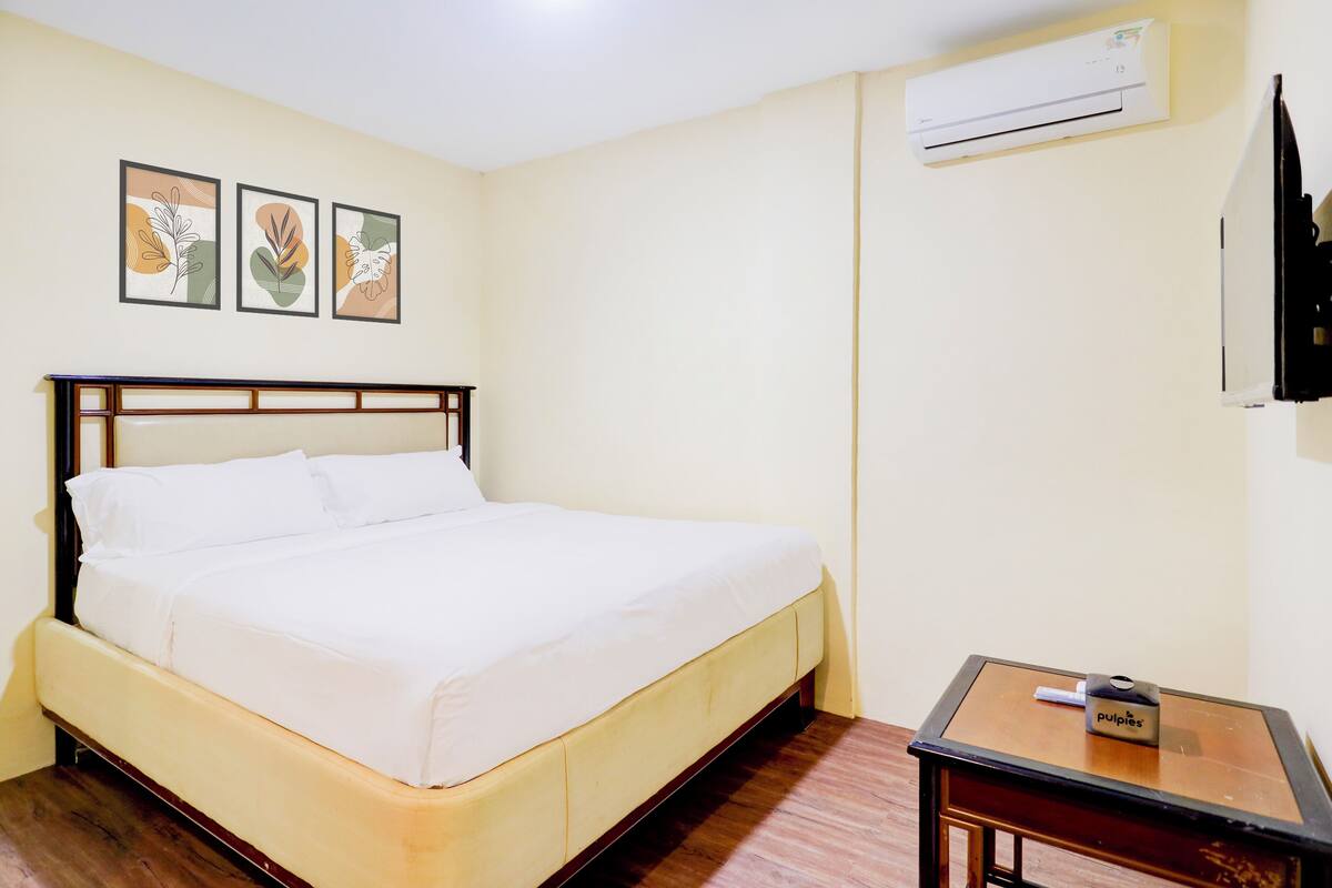 Una cama bien hecha en una habitación con cabecera de madera, una pequeña mesa de madera con una caja etiquetada como “pulpite” y un equipo de aire acondicionado en la pared.