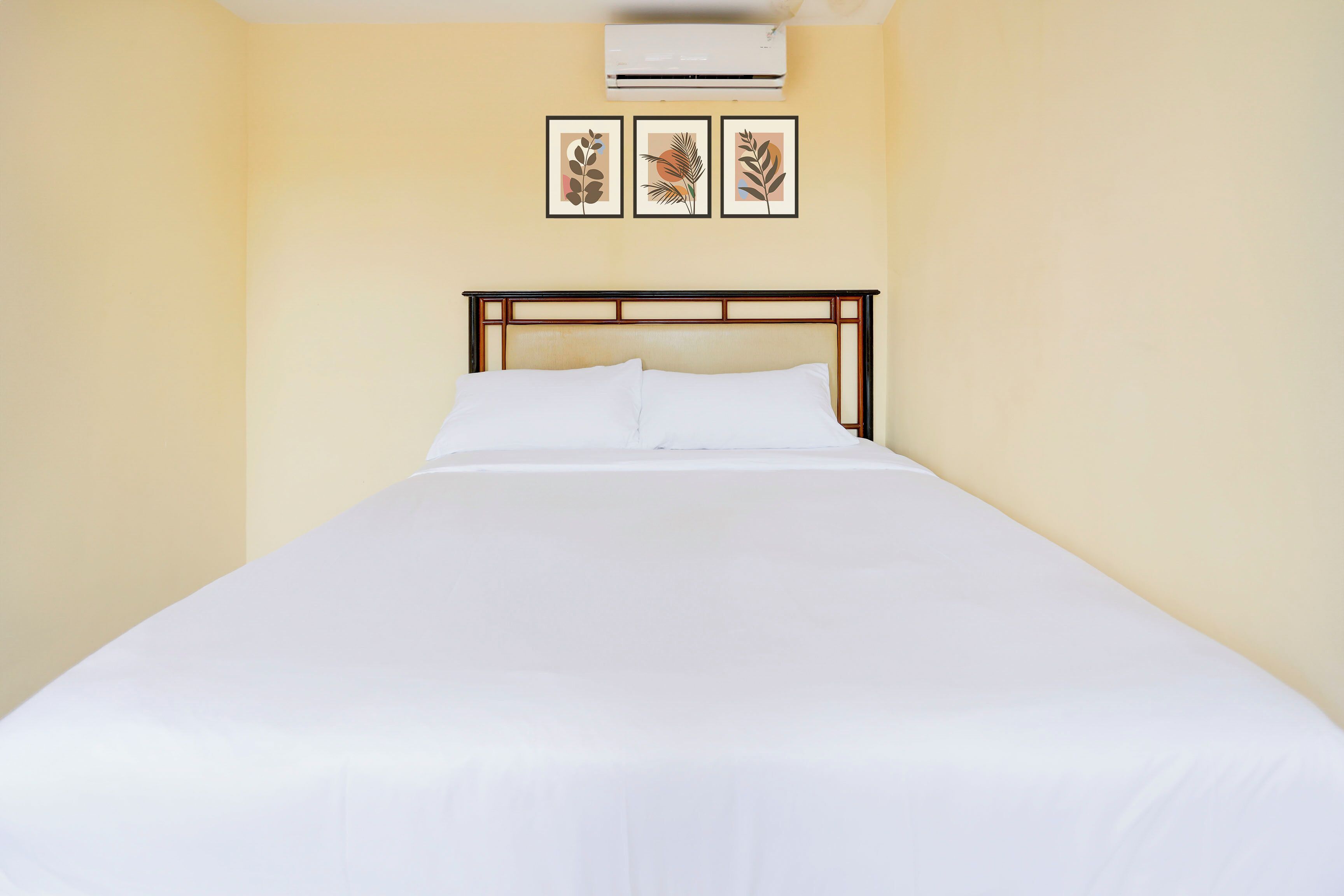 Una cama bien hecha en una habitación con una pared amarilla lisa y una obra de arte enmarcada sobre la cabecera.
