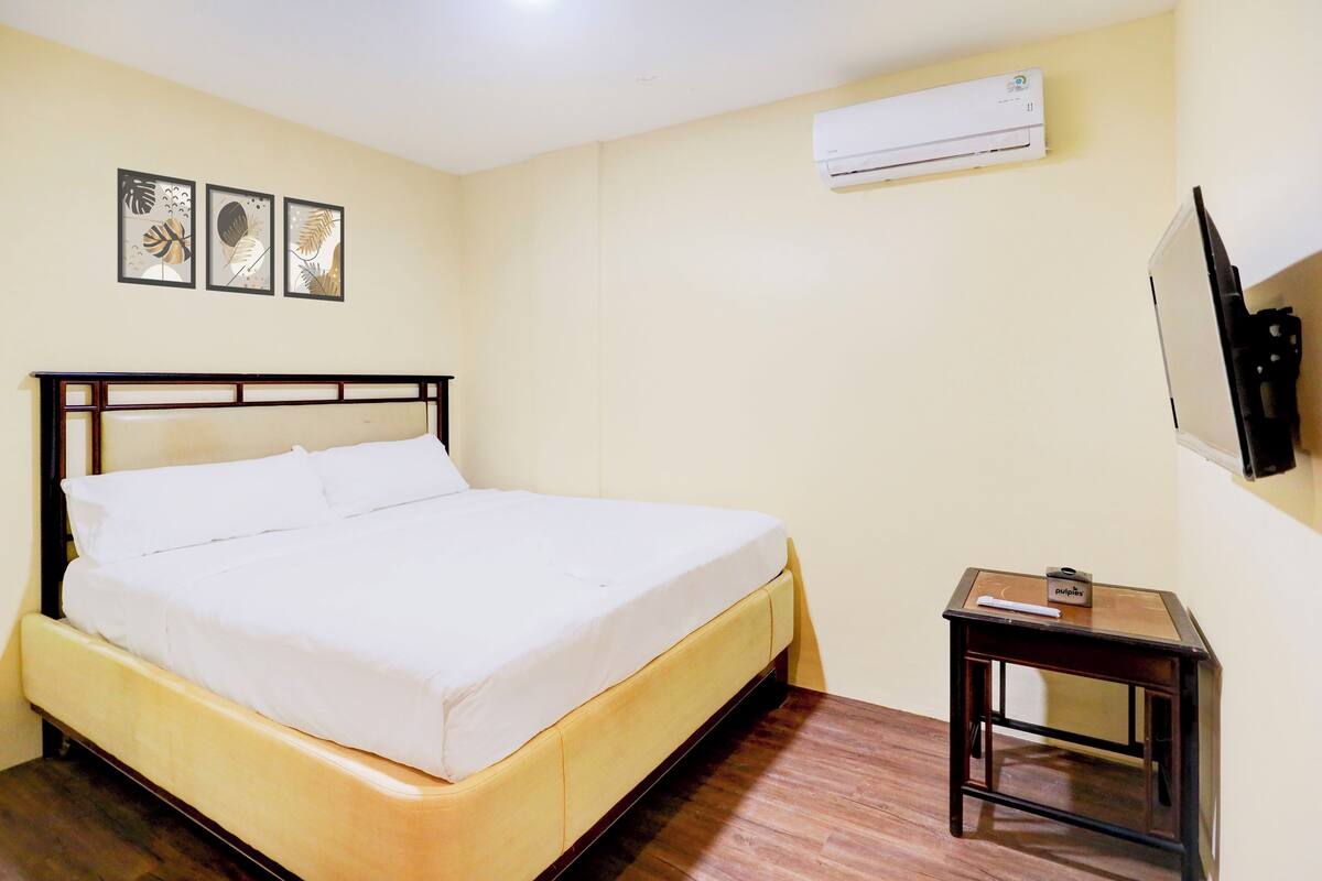 Una cama bien hecha con sábanas blancas, una mesita de noche con un libro y un equipo de aire acondicionado en la pared.