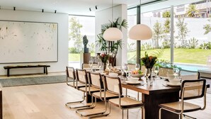 A modern dining area with a long table set for six, surrounded by wicker chairs, with a view of a garden and a whiteboard on the wall.