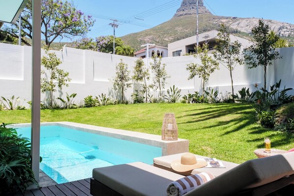 A pool area with lounge chairs, a small table, and a drink. There is a white wall and a mountain in the background.