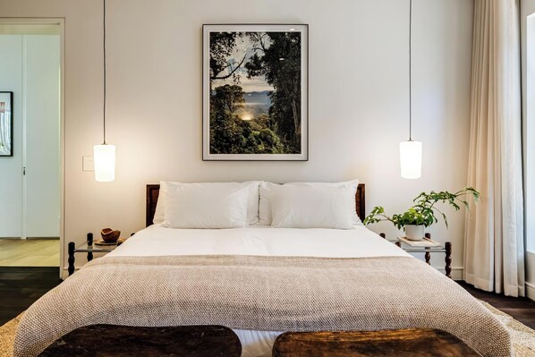 A neatly made bed with white linens, a large framed landscape painting above it, and two bedside lamps.