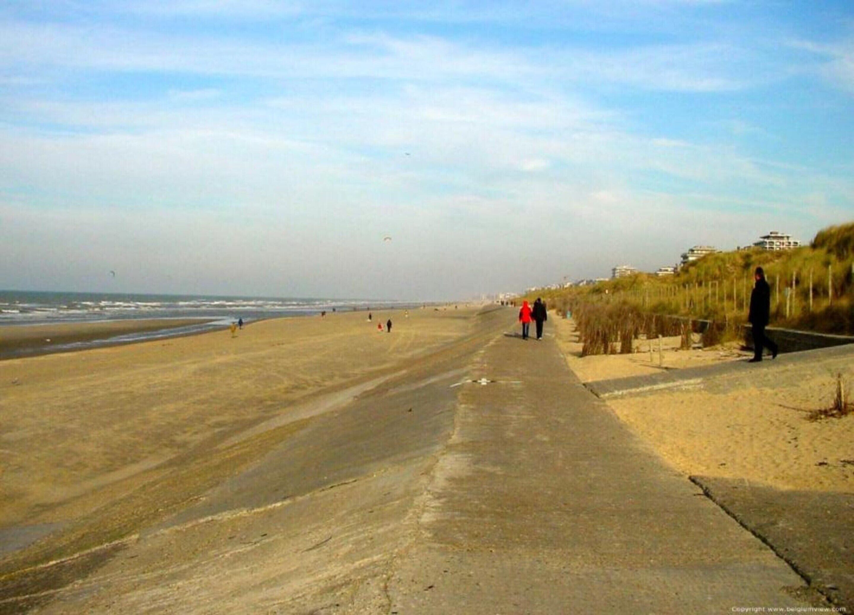 A beach with a concrete walkway, people walking, and buildings in the background.