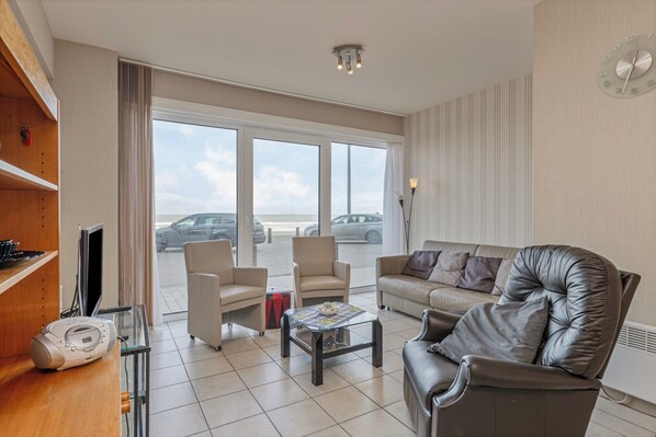 A spacious living room with a leather sofa, armchairs, and a coffee table. A large window offers a view of parked cars outside. - Apartment in Blankenberge Near Seafront (Blankenberge)