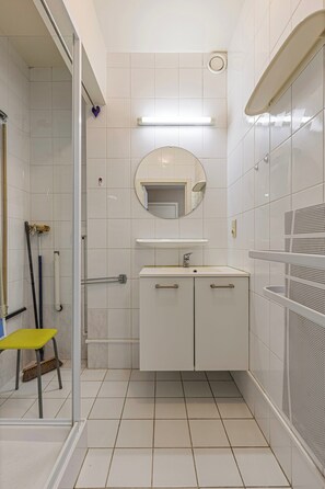 A bathroom with white tiles, a glass shower enclosure, a yellow stool, a mirror, and a sink with cabinets. - Apartment in Blankenberge Near Seafront (Blankenberge)