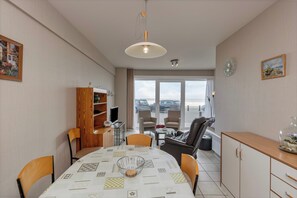 A dining area with a round table, chairs, a wooden cabinet, and a view through a glass door to the outside. - Apartment in Blankenberge Near Seafront (Blankenberge)