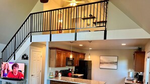 A spacious living area with a large TV, a staircase leading to an upper level, a kitchen with wooden cabinets, and a dining area with bar stools. - Rock Haven - Prescott Cabin Rentals (Prescott)