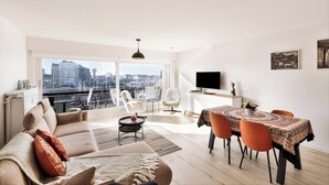 Ein modernes Wohnzimmer mit Sofa, Esstisch, Stühlen, Fernseher und einem Balkon mit Blick auf die Stadt.