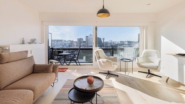 Ein modernes Wohnzimmer mit einer Couch, einem Couchtisch und zwei Sesseln. Ein großes Fenster bietet Blick auf die Stadt.
