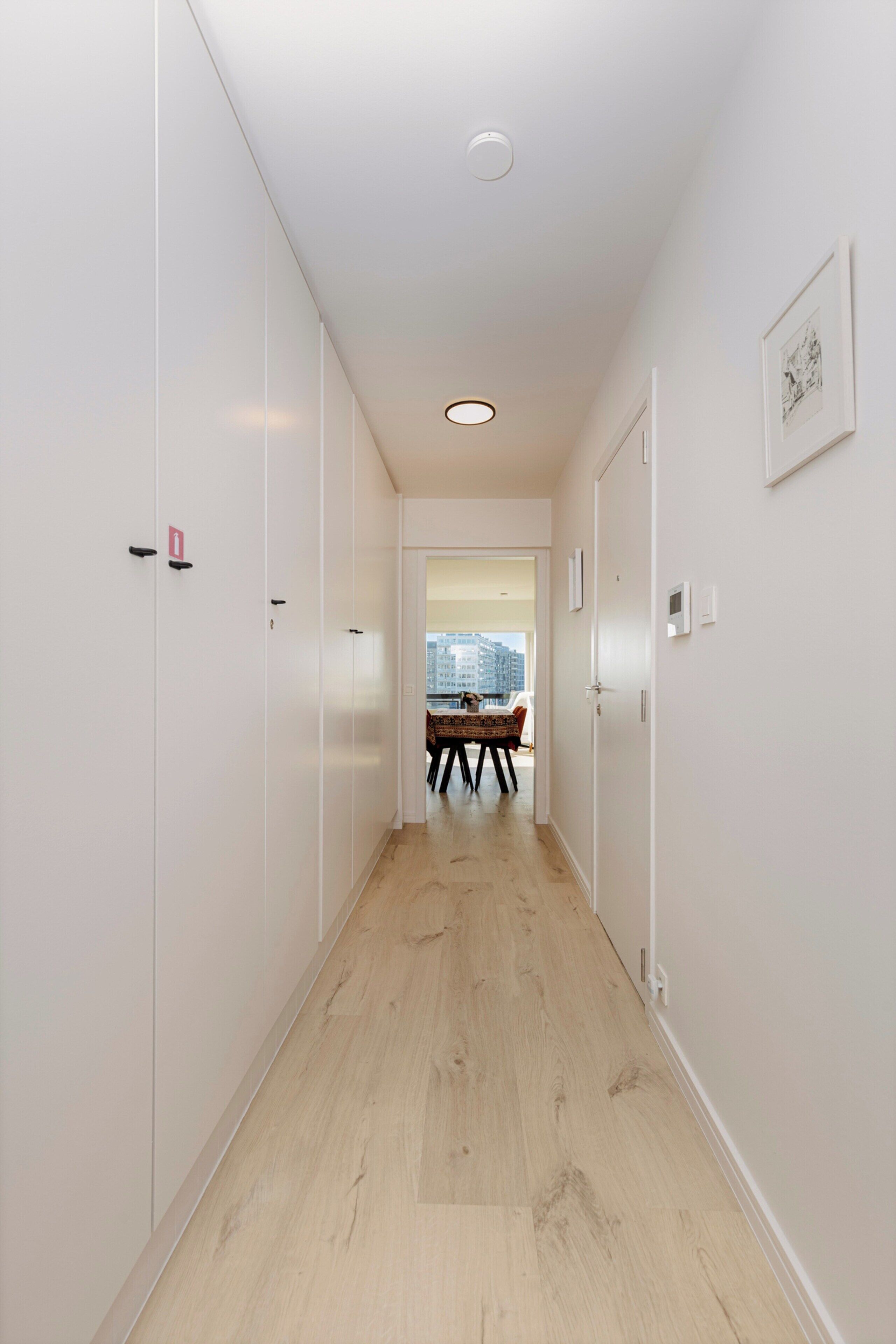 A hallway with white walls, wooden flooring, and a view of a dining area with a table and chairs.