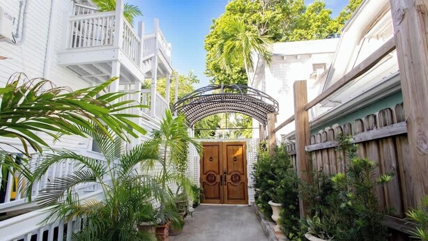 A narrow pathway leading to a wooden door with a metal gate, surrounded by lush greenery and white buildings with balconies. - Sailfish Suite on Duval (Key West)