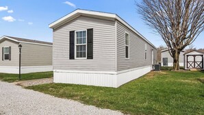 Une maison mobile de plain-pied, dotée d’un porche à l’avant, d’un petit garage attenant et d’un arbre en arrière-plan.