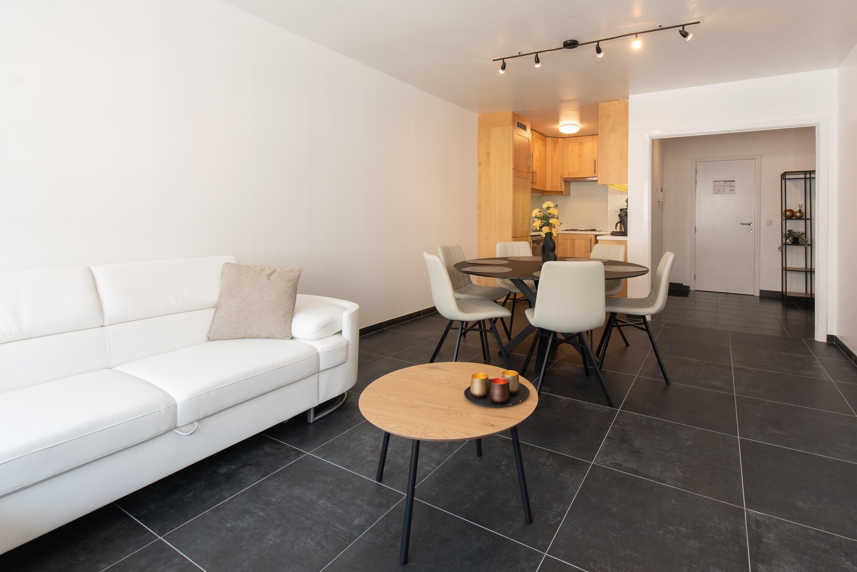 A modern living room with a white sofa, a round wooden coffee table, and a dining area with chairs and a table.