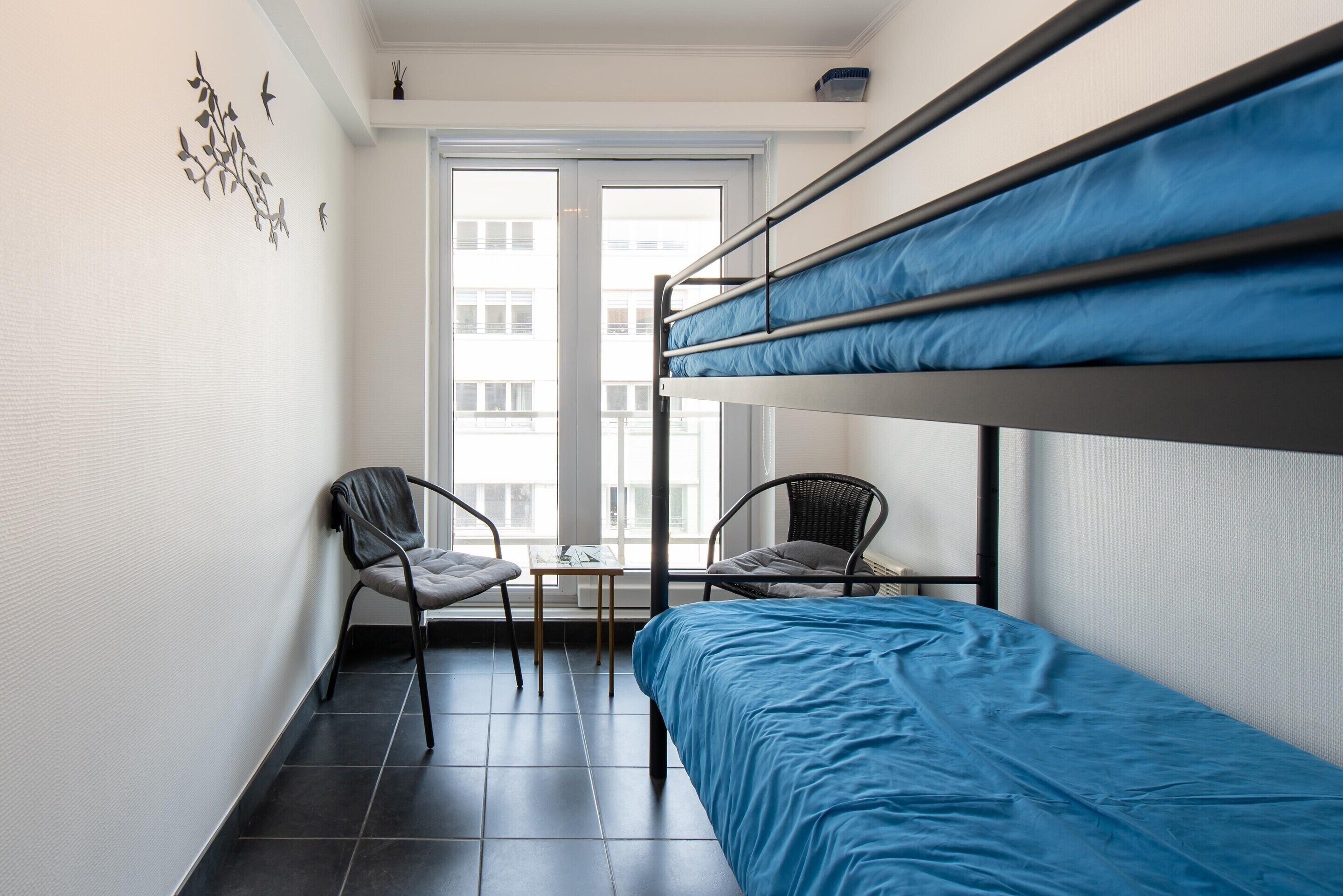 Quarto pequeno e moderno com beliches, uma cadeira, uma mesa e uma janela com vista para edifícios.