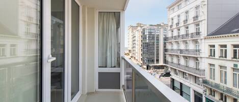 A balcony with a view of adjacent buildings, a window with a curtain, and a glass railing.