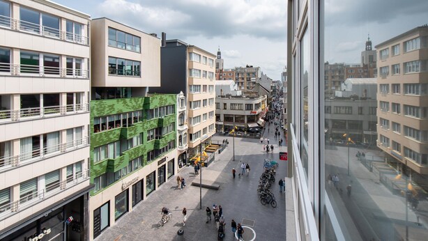 A multi-story building with a ground floor featuring a café and seating area, surrounded by other buildings and a busy street. - Apartment in Ostend Near Shopping Street (Ostend)