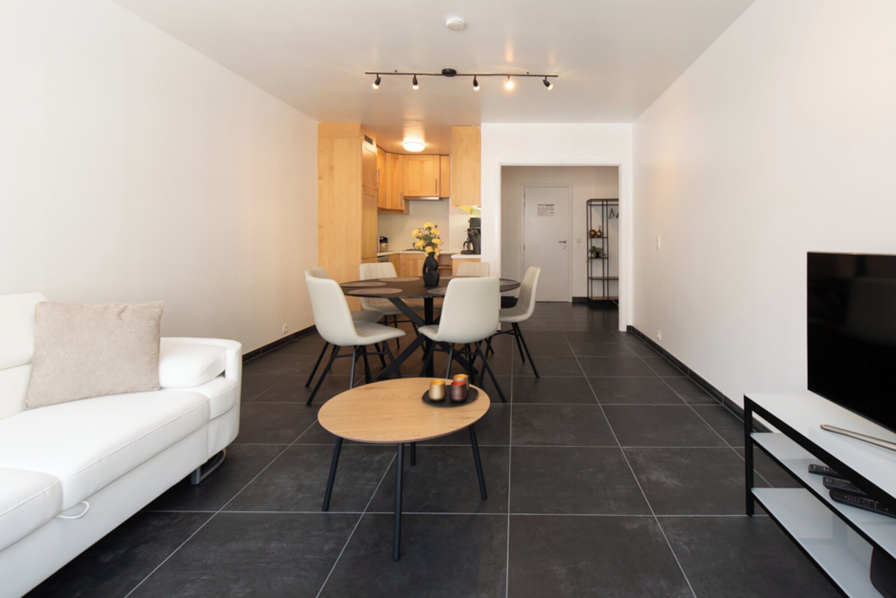 A modern living room with a white sofa, a round coffee table, and a flat-screen TV. There is a dining area with a wooden table and chairs. The room has dark tiled flooring and white walls.