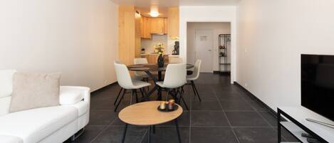 A modern living room with a white sofa, a round coffee table, and a flat-screen TV. There is a dining area with a wooden table and chairs. The room has dark tiled flooring and white walls.