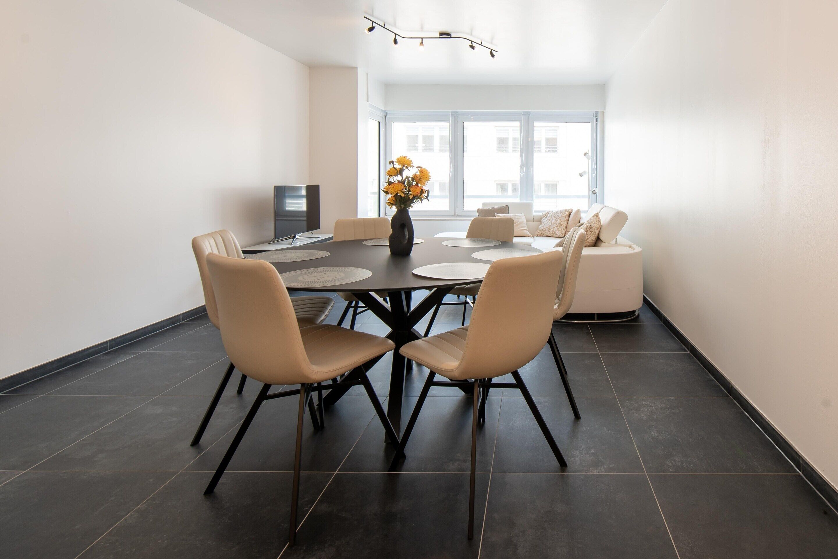 A modern dining area with a round table, beige chairs, a vase with flowers, and a sofa in the background.