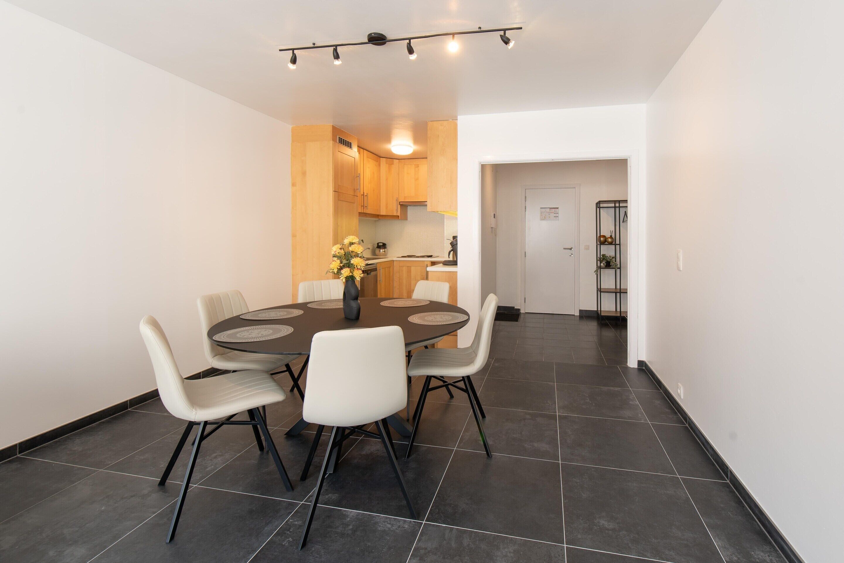A modern dining area with a round table and six chairs, a kitchen in the background, and a hallway leading to a door.