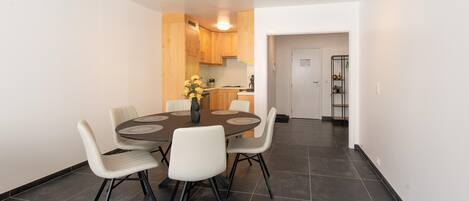 A modern dining area with a round table and six chairs, a kitchen in the background, and a hallway leading to a door.