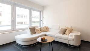 A modern living room with a white sectional sofa, a small round coffee table, and a large window providing natural light. - Apartment in Ostend Near Shopping Street (Ostend)