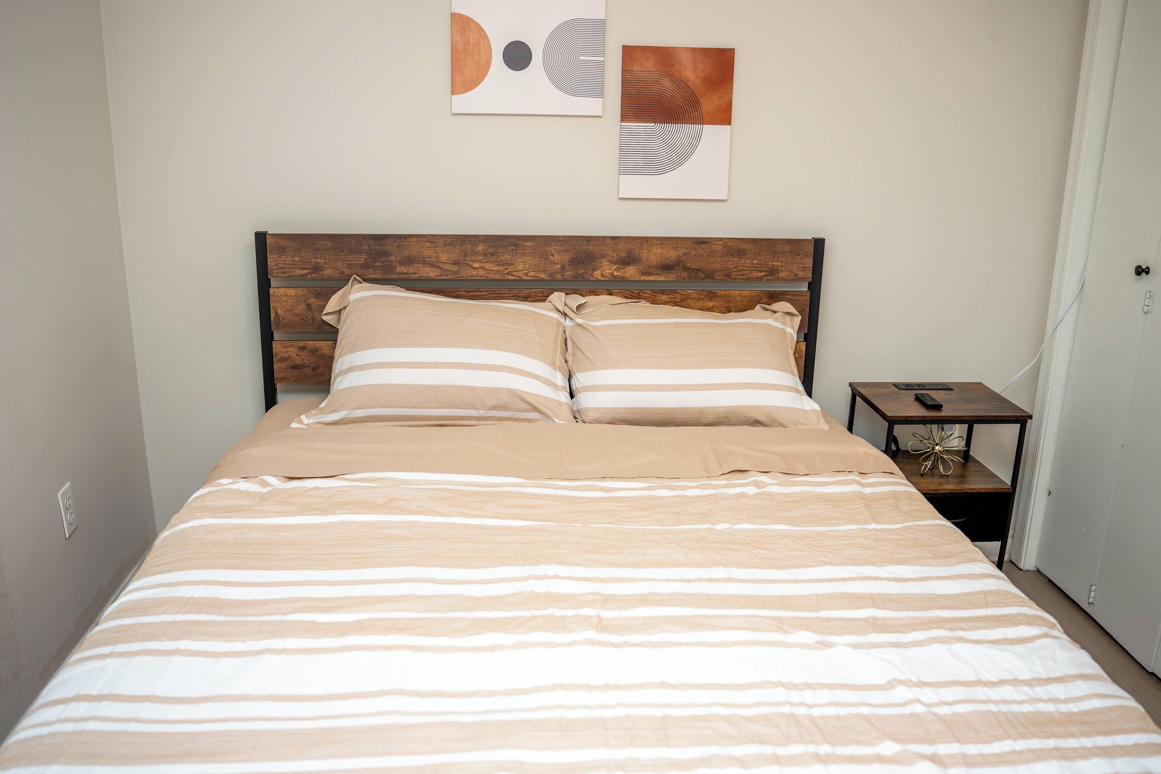 Un lit avec une literie à rayures, une tête de lit en bois et deux œuvres d’art abstraites accrochées au mur.