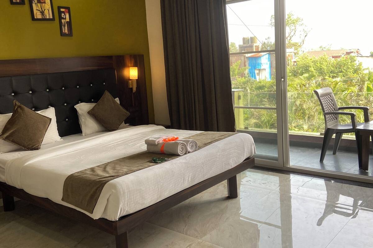 A hotel room with a large bed, a ceiling fan, a view of greenery outside, and a balcony with a chair.