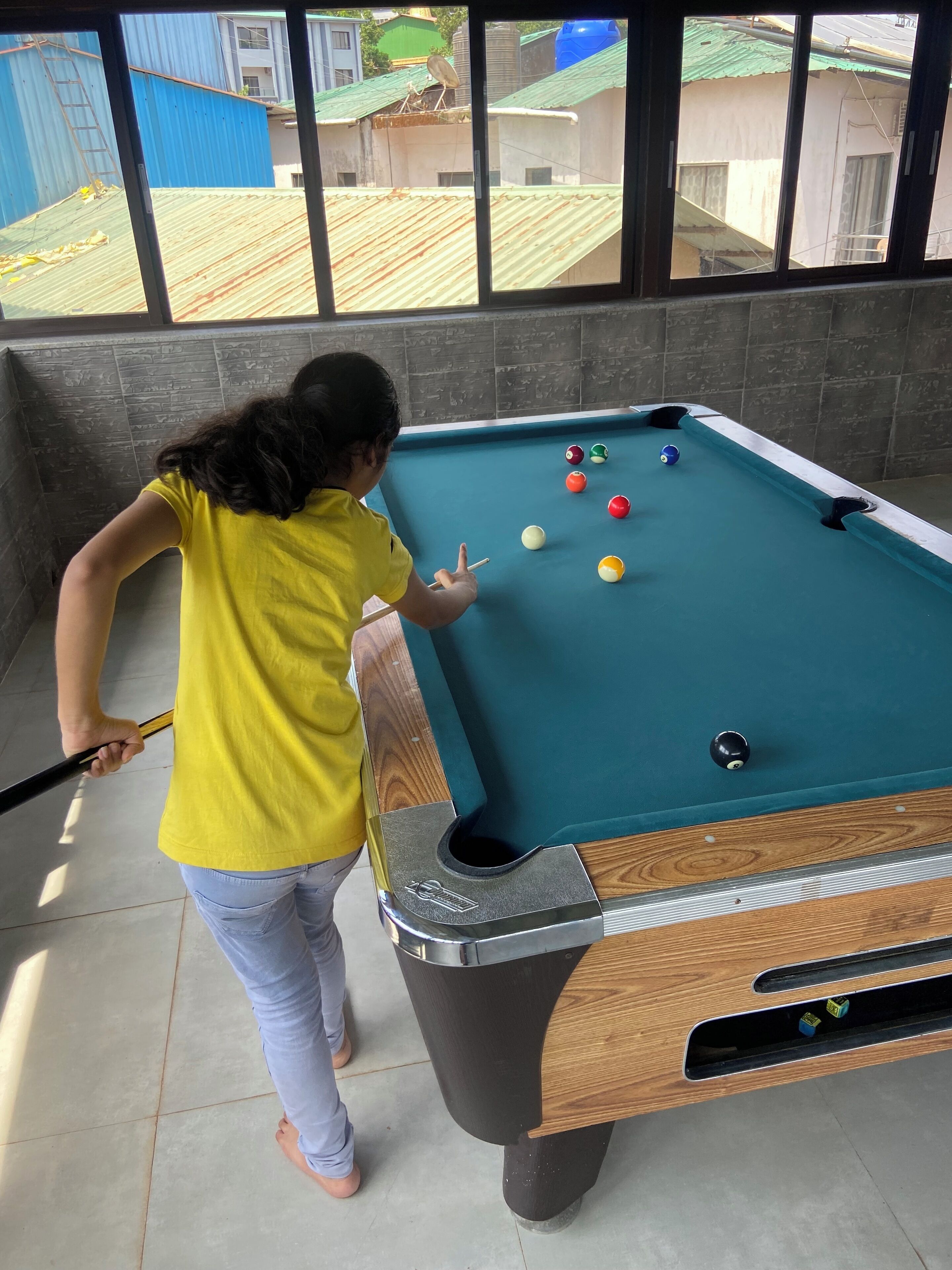 A pool table with balls and a cue stick, a window with a view of buildings, and a person in a yellow shirt.