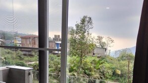 A room with a large window overlooking a green area with trees and a building. There is a table and a chair inside the room. - Hotel Om Palace (Mahabaleshwar)
