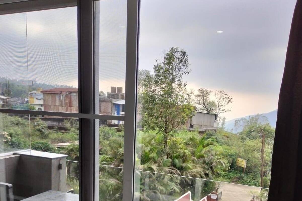 A room with a large window overlooking a green area with trees and a building. There is a table and a chair inside the room.