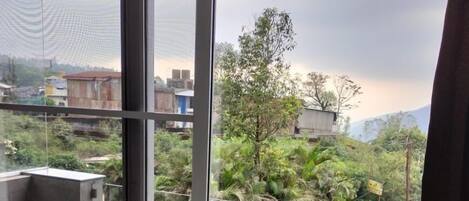 Habitación con un ventanal grande que da a un área verde con árboles y un edificio. Hay una mesa y una silla dentro de la habitación.