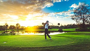 Eine Person spielt Golf an einem sonnigen Tag bei blauem Himmel, im Hintergrund ist ein Gewässer zu sehen.