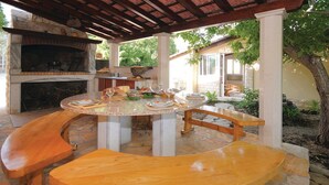 An outdoor patio with a stone fireplace, wooden table, and benches. - Picturesque Escape Holiday Home With Pool and Sauna (Kastela)