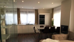 Een moderne woonkamer met een bank, een glazen scheiding, een eethoek met een tafel en stoelen, en een keuken met een fornuis en spoelbak.
