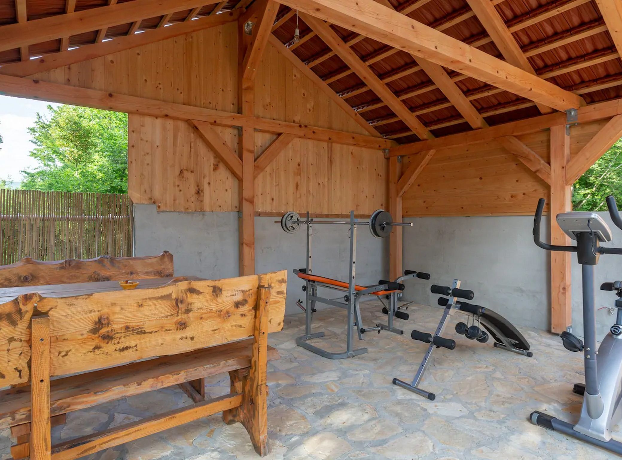 A covered outdoor gym with a bench, weight bench, barbell, dumbbells, and an elliptical machine.
