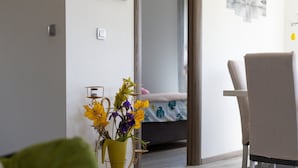 Una sala moderna con un jarrón amarillo lleno de flores, un sofá gris y una silla. Se ve una cama con cubrecama floral al fondo.