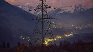 A power line tower with multiple cross-arms and insulators, set against a mountainous backdrop with snow-capped peaks and a town with lights in the distance. - Hygge Home Manali (Manali)