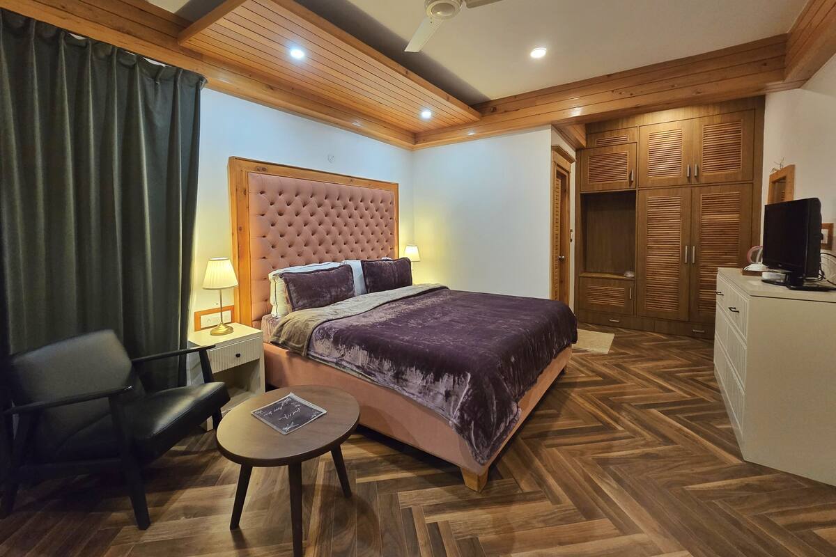 A hotel room with a large bed, a desk, a chair, a ceiling fan, and wooden flooring.