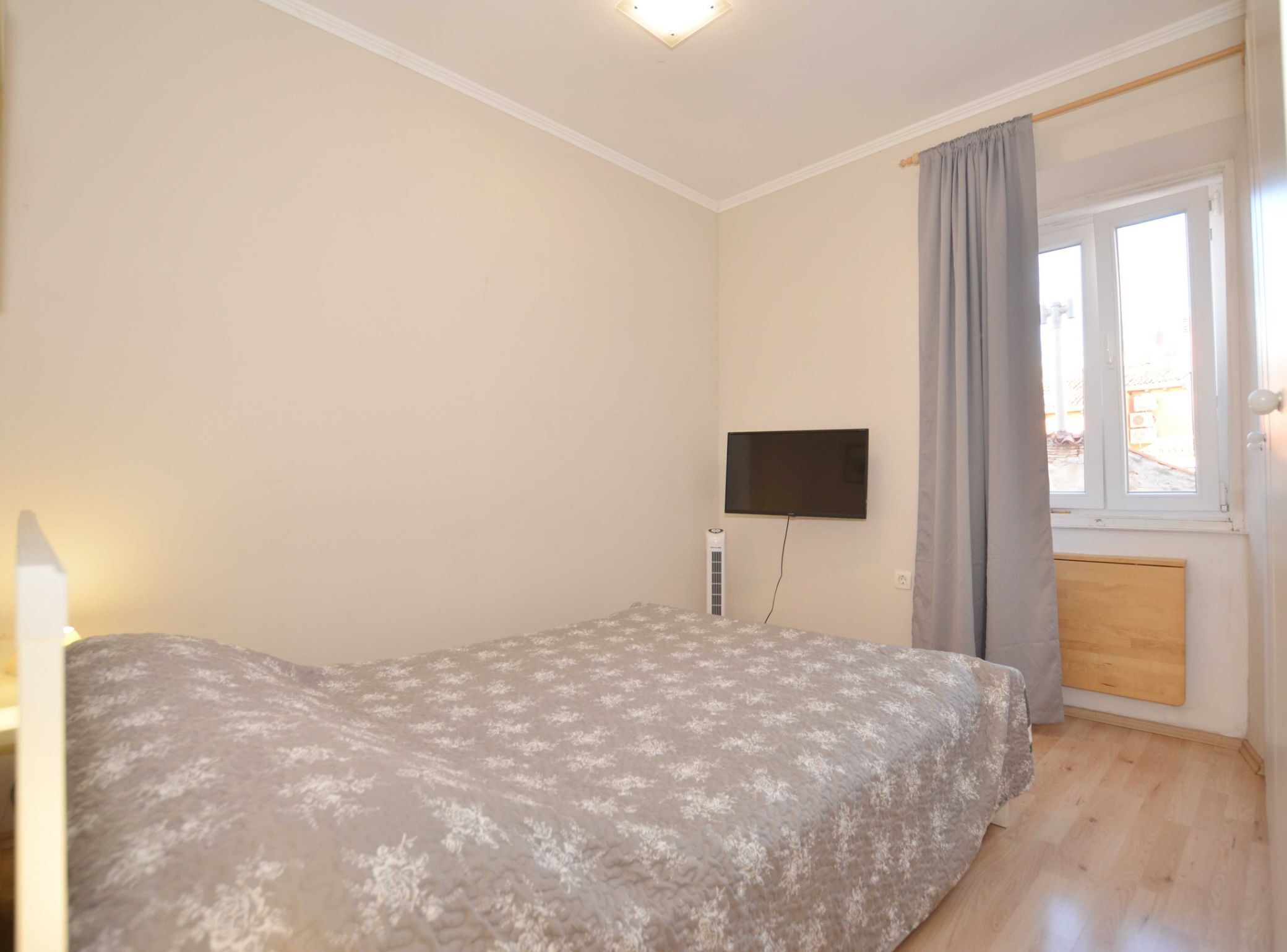 A bedroom with a bed, a TV mounted on the wall, a window with curtains, and a wooden door.