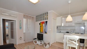 A modern living room with a sofa, a glass coffee table, a TV, and a small white table. There is a kitchen area with a stove, sink, and cabinets. - Seaside Charm Apartment in Rovinj (Rovinj)