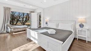 A bedroom with a large bed, a chaise lounge, and a view of trees and houses through the window. - Waterfront Retreat with Private Dock and Fire Pit  |  Sleeps 8, Outdoor Dining, Spa-Style Primary, Chef’s Kitchen w/ Dual Dishwashers, Gas Fireplace, River Views, Bright Open Layout, Central A/C, Perfect Coastal Escape for Families, 24 Hr Hospitality (Falmouth)