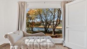 A bedroom with a large window, a tufted sofa, and a view of a lake and trees. - Waterfront Retreat with Private Dock and Fire Pit  |  Sleeps 8, Outdoor Dining, Spa-Style Primary, Chef’s Kitchen w/ Dual Dishwashers, Gas Fireplace, River Views, Bright Open Layout, Central A/C, Perfect Coastal Escape for Families, 24 Hr Hospitality (Falmouth)